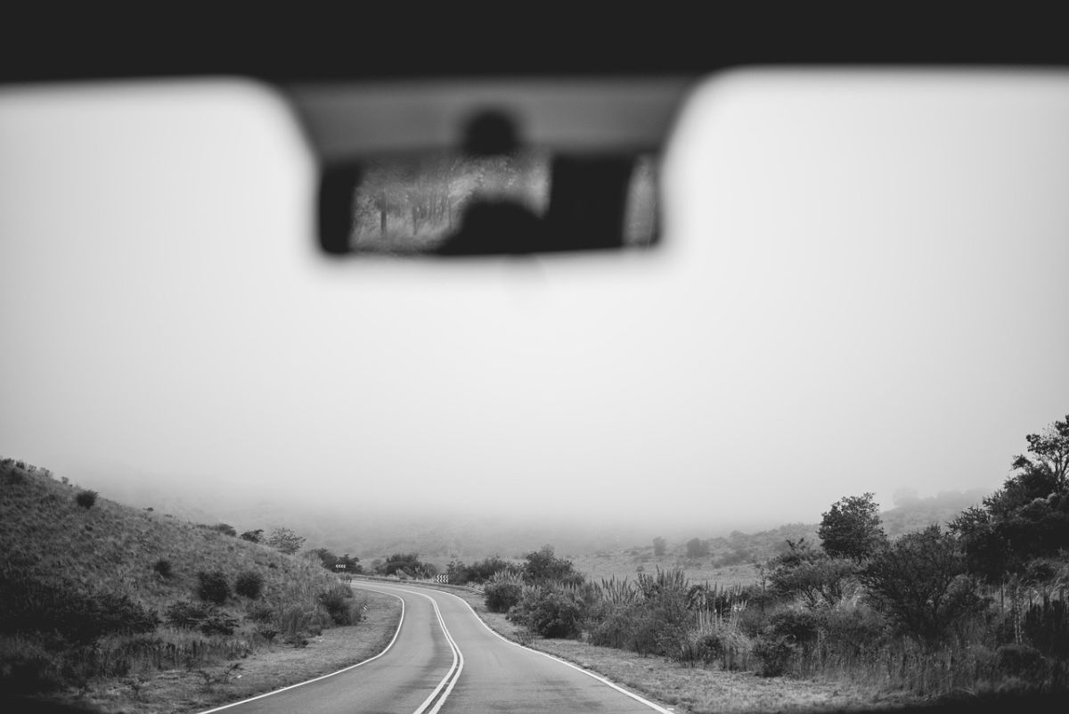 Blick in den Rückspiegel eines Autos. Vor uns in schwarz weiß eine leicht engenebelte Straße und Landschaft.
